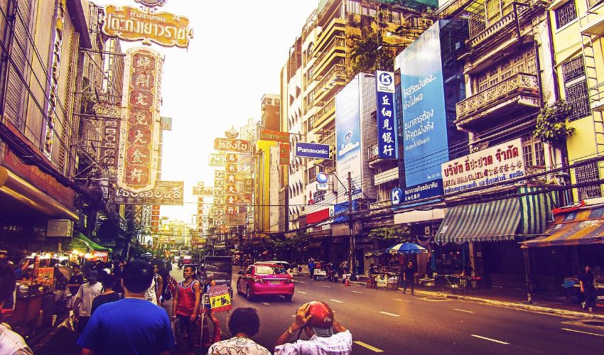 Eine Seitenstraße in Bangkok mit vielen Schildern auf den Häusern und vielen Menschen links und rechts von der Straße.