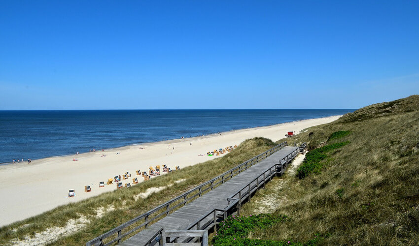 Ein langer Sandstrand entlang der Küste mit einem Holzsteg als Zugang.