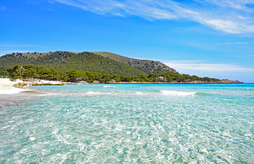 Türkises, flaches Meer mit Sandstrand und links daneben ein mit Bäumen bewachsener Hügel.