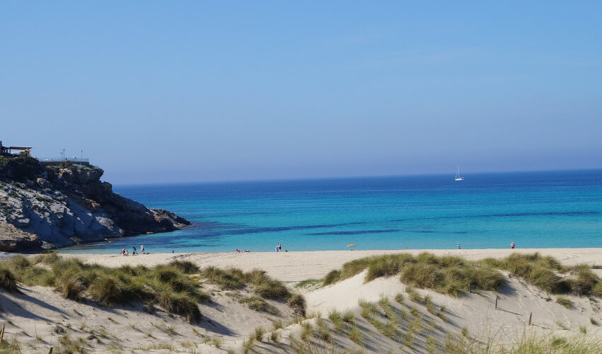 Ein weißer Sandstrand dahinter Sand mit Dünen und ein weiter Blick auf das blaue Meer.