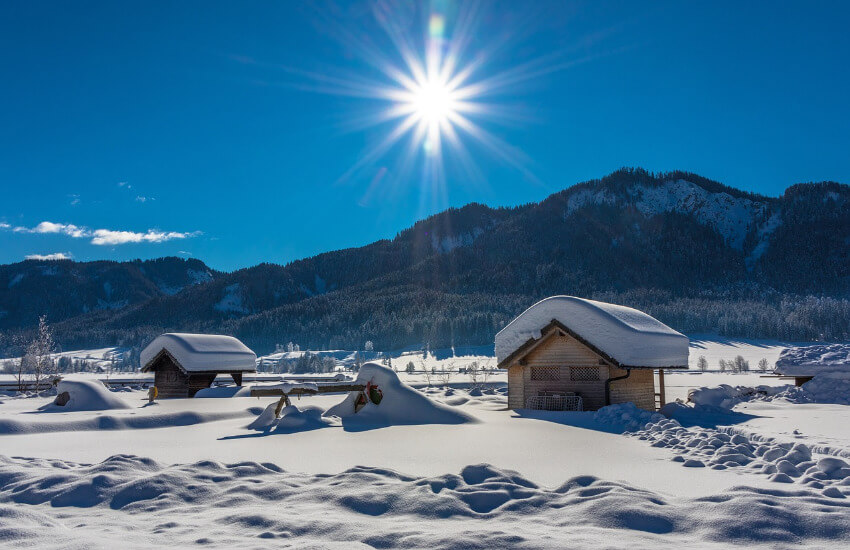 Zwei kleine Holzhütten mit einer dicken Schneedecke auf dem Dach in einer Winterlandschaft unter Sonnenschein.