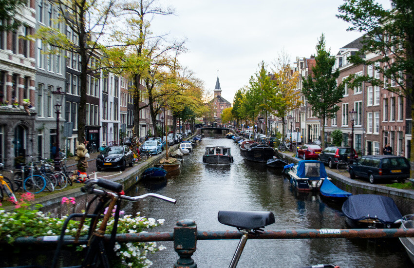 Ein Kanal/Gracht in Amsterdam, wo links und recht Boote parken und in der Mitte des Kanals ein Boot durchfährt.