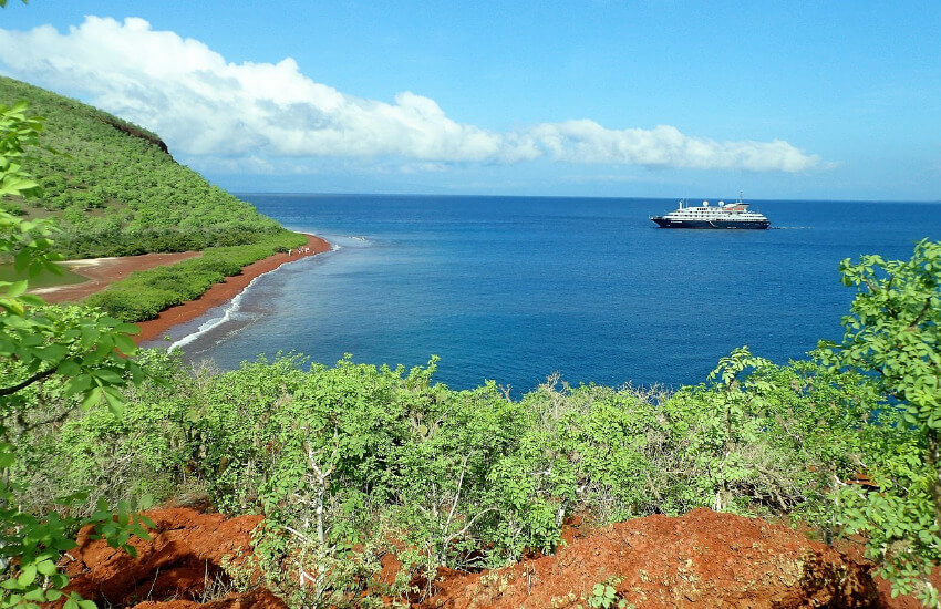 Eine mit Sträuchern bewachsene Galapagos Insel. Ein kleines Kreuzfahrtschiff ankert kurz vor der Insel auf dem Meer.