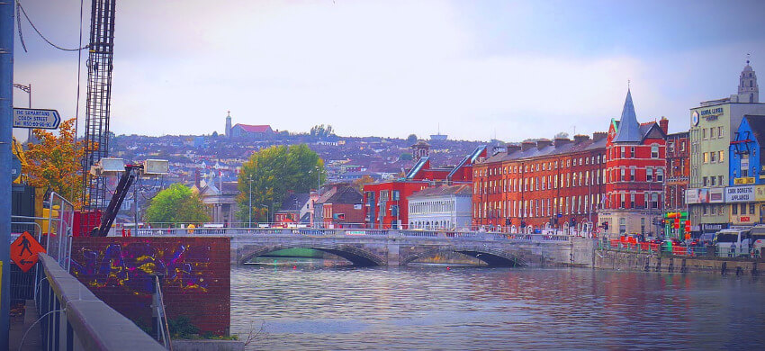 Bunte Häuserzeile am Ufer des Flusses Lee in Cork, Irland, mit Spiegelungen im Wasser bei klarem Wetter.