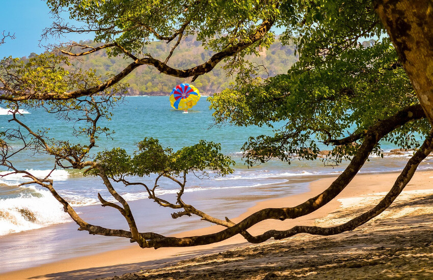 Tropische Küstenlandschaft in Manuel Antonio, Costa Rica: Eine von dichtem grünem Regenwald gesäumte Bucht trifft auf den türkisblauen Pazifik unter einem klaren Himmel. Die bewaldeten Hügel reichen bis an die Küste und bilden eine malerische, naturbelassene Kulisse.